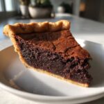 A close-up slice of rich, dark chocolate chess pie resting on a white plate, showing the cracked top and buttery crust.