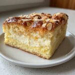 A close-up of a square slice of bee sting cake showing layers of sponge, cream filling, and a caramelized almond topping.