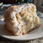 Close-up of a moist slice of coconut cake covered in white frosting and toasted coconut flakes.