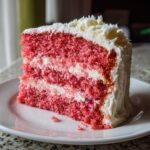 A tall slice of vibrant pink layer Cherry cake filled and frosted with white cream cheese frosting, served on a white plate.