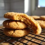 A stack of three soft peanut butter cookies, one broken in half to show the chewy interior, resting on a cooling rack.