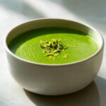 A close-up of vibrant green asparagus soup in a white bowl, topped with bright yellow lemon zest.