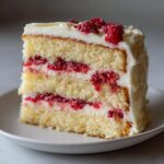 A close-up slice of white chocolate raspberry cake showing three layers of vanilla sponge, raspberry filling, and white frosting.