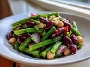 A vibrant close-up of a zesty bean salad featuring green beans, dark red kidney beans, chickpeas, and red onion slices.
