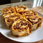Six baked taco pinwheels filled with seasoned ground beef and melted cheese, served on a white plate.