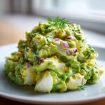 A mound of creamy avocado egg salad featuring chunks of hard-boiled egg and diced red onion, garnished with fresh dill.