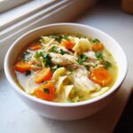 Close-up of a white bowl filled with steaming chicken noodle soup, featuring shredded chicken, bright orange carrots, and parsley.