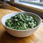 A white bowl filled with rich, homemade creamed spinach sitting on a wooden surface.