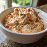 A close-up of creamy, orange-hued boat dip mixed with red peppers and green herbs in a white bowl.