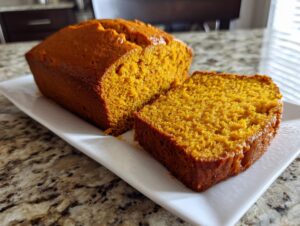 A loaf of moist, orange-colored easy pumpkin bread with one thick slice cut and displayed next to it on a white platter.