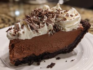 A rich slice of french silk pie featuring a dark chocolate crust, thick chocolate mousse filling, and topped with whipped cream and chocolate shavings.