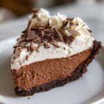 A rich slice of french silk pie showing the dark chocolate crust, creamy filling, and whipped cream topped with chocolate shavings.