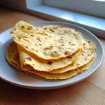 A stack of freshly made, warm yellow corn tortillas with light brown char marks on a light gray plate.