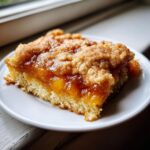 A close-up of a square slice of peach cobbler with a thick, gooey peach filling and cinnamon crumble topping.