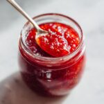 Close-up of a spoonful of bright red strawberry freezer jam being lifted from a glass jar.