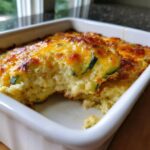 A scoop has been taken from the Addictive Zucchini Casserole, revealing a creamy interior and a golden, cheesy baked top.