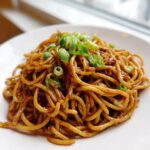A close-up shot of glossy, saucy garlic noodles topped with fresh sliced green onions on a white plate.