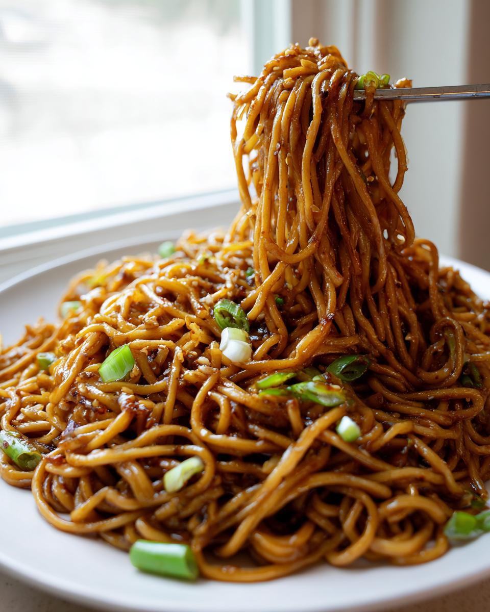 A large serving of glossy, brown garlic noodles being lifted high with chopsticks from a white plate.