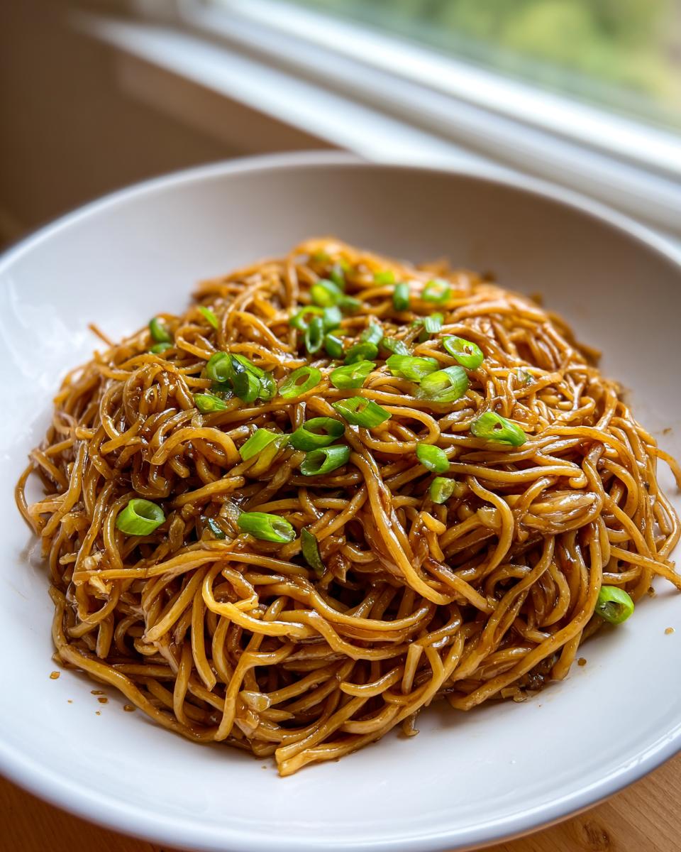A close-up of a white bowl filled with glossy, saucy garlic noodles topped with fresh sliced green onions.