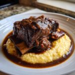 A close-up of a tender braised short ribs portion served over yellow creamy polenta, drenched in rich dark sauce.