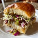 A close-up of a delicious grinder sliders on a white plate, featuring melted cheese, shredded lettuce, red onion, and green relish.