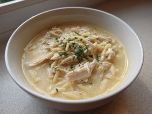 A close-up of a white bowl filled with Classic Greek Lemon Chicken Soup, featuring shredded chicken, orzo pasta, and a dill garnish.