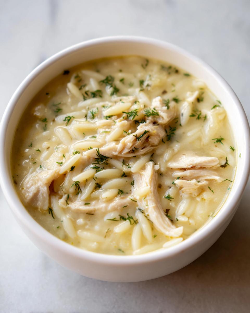 Close-up of a white bowl filled with Classic Greek Lemon Chicken Soup featuring shredded chicken, orzo pasta, and fresh dill.