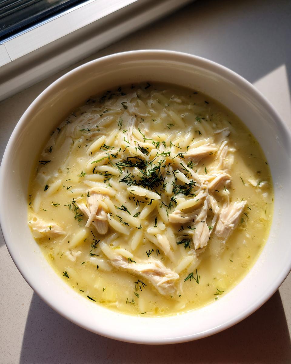 A close-up overhead view of a white bowl filled with Classic Greek Lemon Chicken Soup, featuring shredded chicken, orzo pasta, and fresh dill.