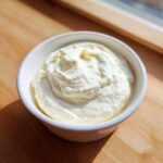 A close-up of creamy, homemade garlic aioli recipe served in a small white bowl on a wooden surface.