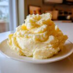 A large, fluffy mound of creamy, yellow-tinted mashed potatoes served on a white plate.