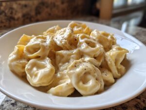 Close-up of a white bowl filled with rich Tortellini Alfredo Recipe, topped with cracked black pepper.