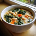 Close-up of a white bowl filled with Easy Tuscan White Bean Soup, featuring white beans, dark green kale, and orange carrots.