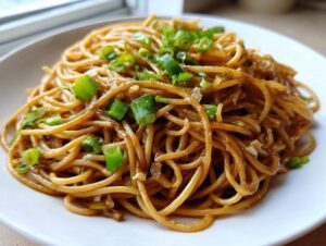 A close-up shot of glossy, saucy garlic noodles topped with fresh chopped green onions served on a white plate.