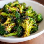Close-up shot of vibrant green Honey Mustard Roasted Broccoli florets coated in a glistening, grainy sauce, served in a white bowl.