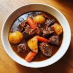 A white bowl filled with rich, dark brown beef stew featuring chunks of tender beef, whole small potatoes, and bright orange carrots.