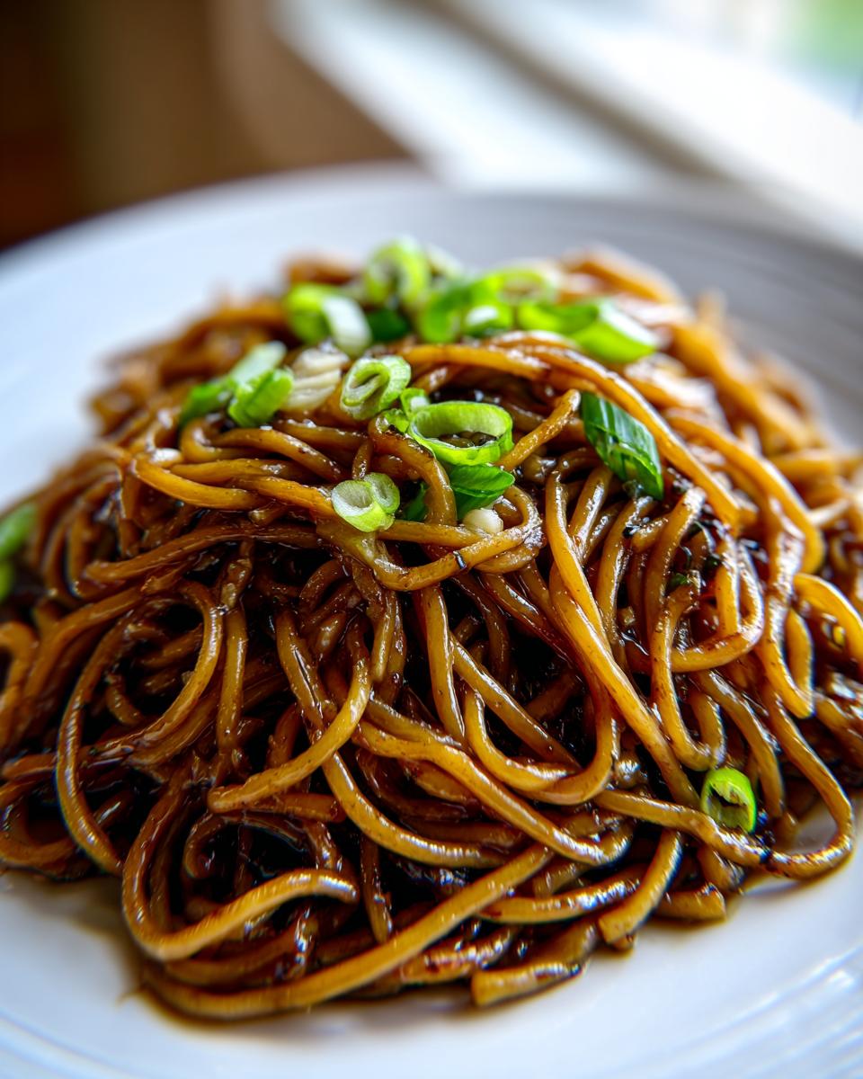 A close-up shot of glossy, dark brown garlic noodles piled high on a white plate and garnished with fresh sliced green onions.