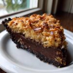 A close-up slice of german chocolate pie featuring a dark chocolate filling and toasted coconut topping on a white plate.