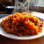 A mound of steaming, vibrant orange-red jollof rice served on a white plate, backlit by natural light.